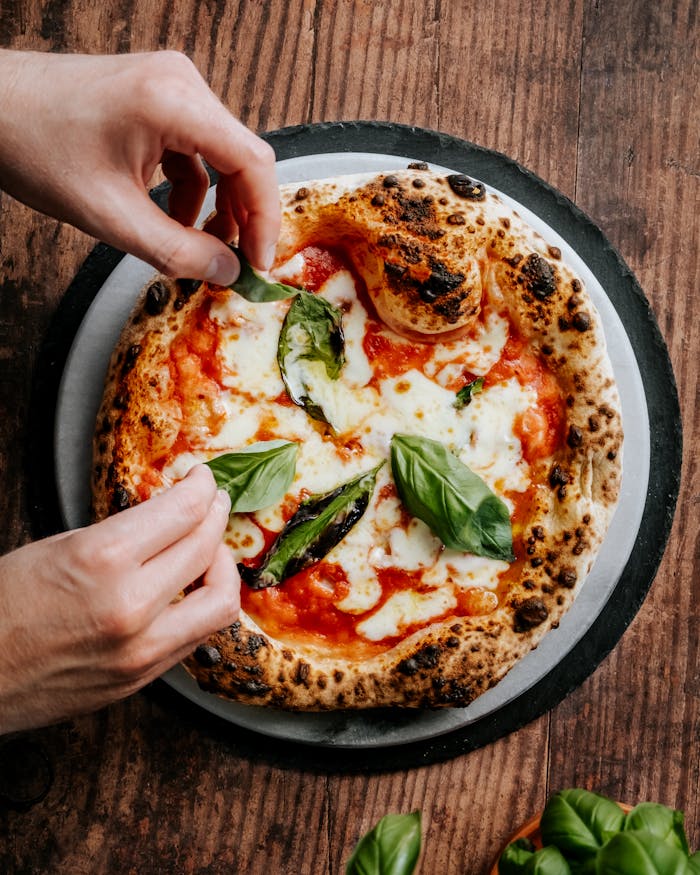 Close-up of hands adding basil to a freshly baked Neapolitan pizza, highlighting its cheesy and delicious appeal.